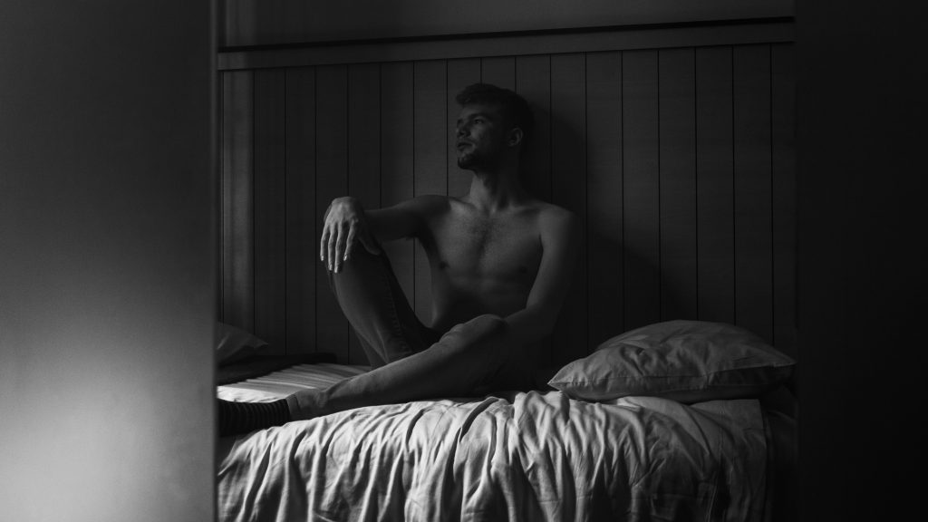 Man sitting on edge of bed in a dim room, reflecting with low energy.